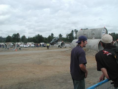 Dark SUV driving over large concrete pipes at an outdoor event.