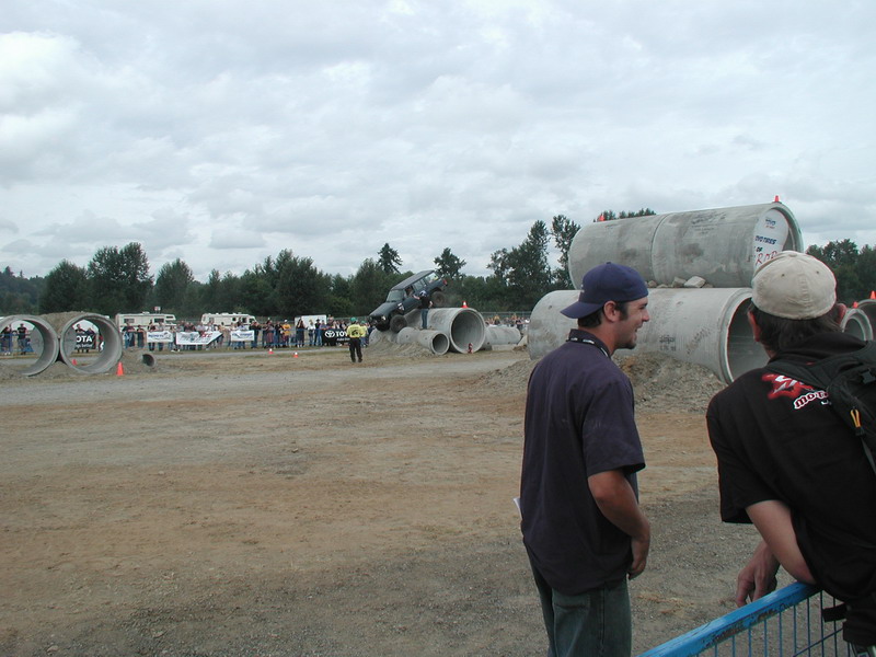 Dark SUV driving over large concrete pipes at an outdoor event.