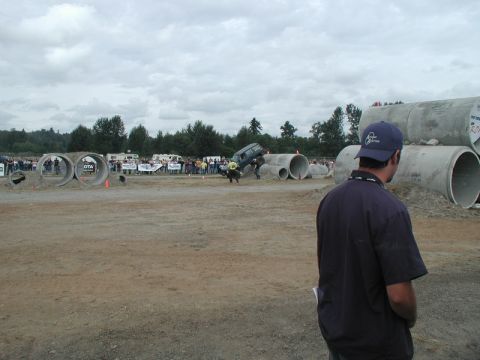 Dark SUV jumping over a concrete pipe obstacle at an outdoor event.