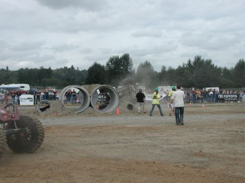 Modified off-road vehicle with large tires going through a concrete pipe obstacle with spectators watching.