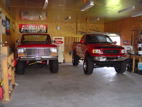 1978 Ford Bronco - Two red Ford trucks, a lifted 1978 Bronco and a newer pickup, are parked inside a garage.