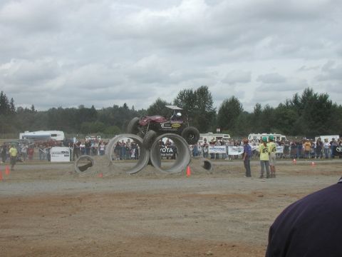 Maroon off-road buggy jumping over concrete pipes at an outdoor event with spectators.