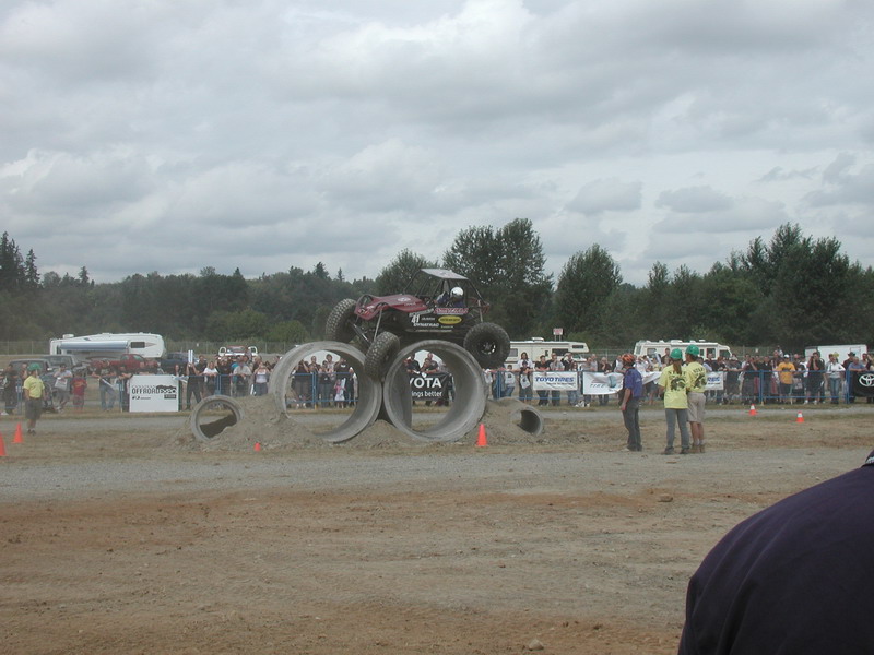 Maroon off-road buggy jumping over concrete pipes at an outdoor event with spectators.