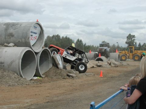Orange and white off-road buggy climbing over concrete pipes at an event.