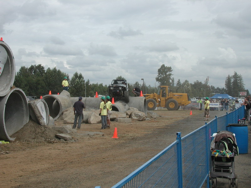 Black off-road vehicle on a pipe and rock obstacle course with a front-end loader and spectators.