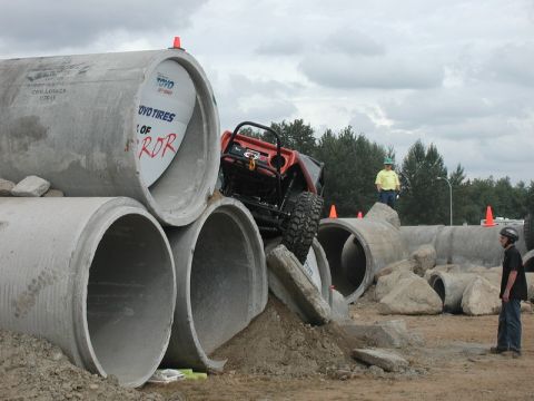Orange off-road vehicle climbing over concrete pipes with a person watching.