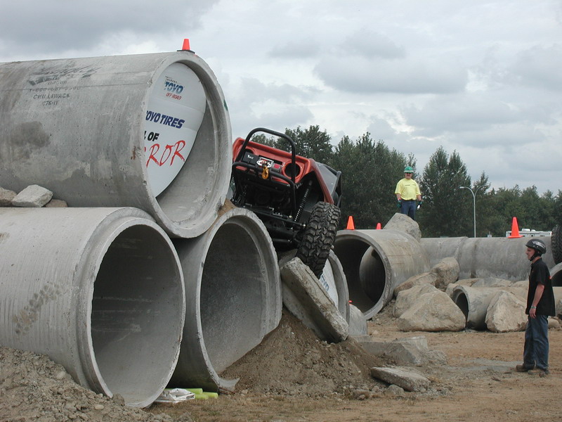 Orange off-road vehicle climbing over concrete pipes with a person watching.