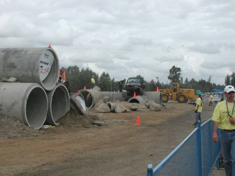 Black off-road vehicle on a pipe obstacle course with a yellow loader and staff nearby.