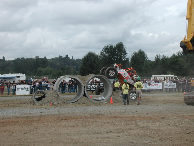 Orange off-road buggy upside down after crashing through concrete pipes, crowd watching.