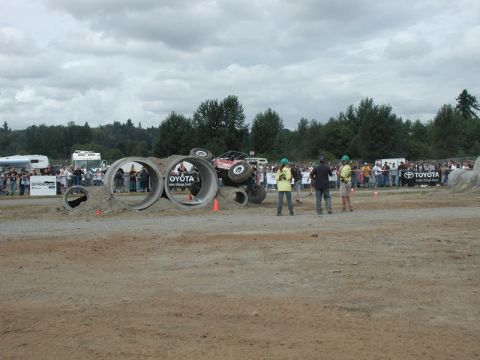 Red off-road vehicle tilted on side going through concrete pipes with spectators watching.