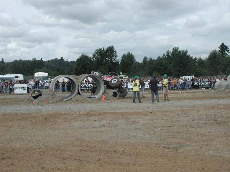 Red off-road vehicle tilted on side going through concrete pipes with spectators watching.