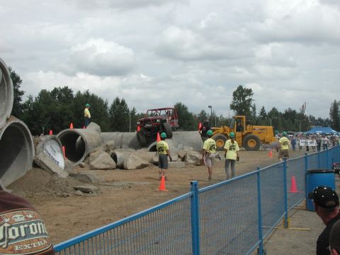 Red off-road buggy climbing over concrete pipes and rocks at an outdoor event.