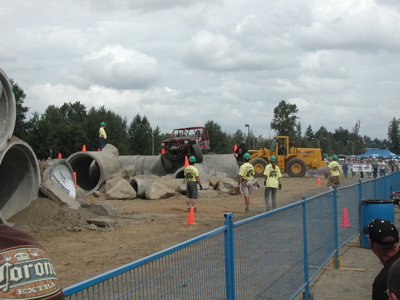 Red off-road buggy climbing over concrete pipes and rocks at an outdoor event.