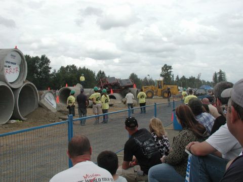 Monster truck on concrete pipes with spectators and a front-end loader.