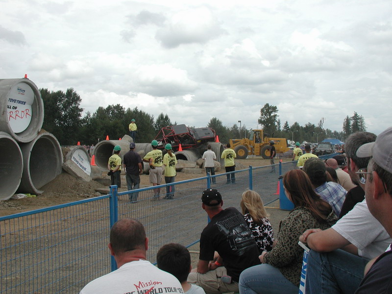 Monster truck on concrete pipes with spectators and a front-end loader.
