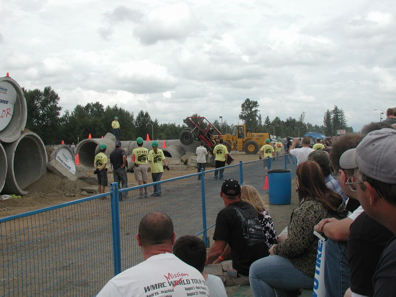 Custom off-road buggy airborne over concrete pipes at an event, with staff and spectators watching.