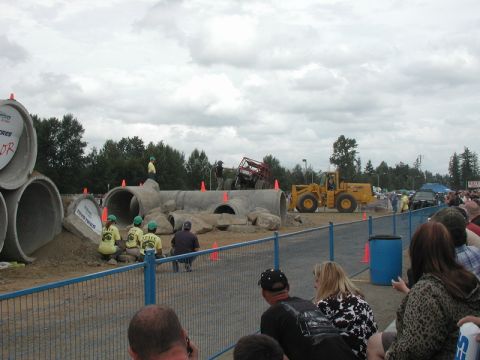 Red off-road vehicle on concrete pipes with yellow loader and spectators.