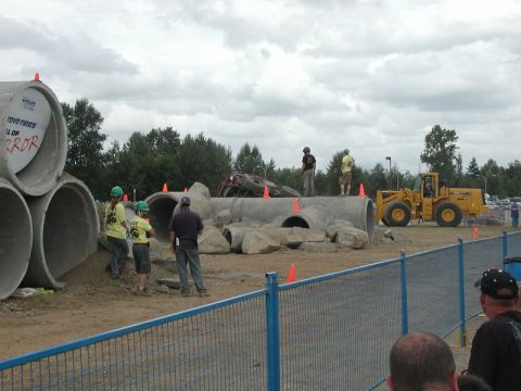 Red off-road vehicle on concrete pipes with yellow loader and observers.