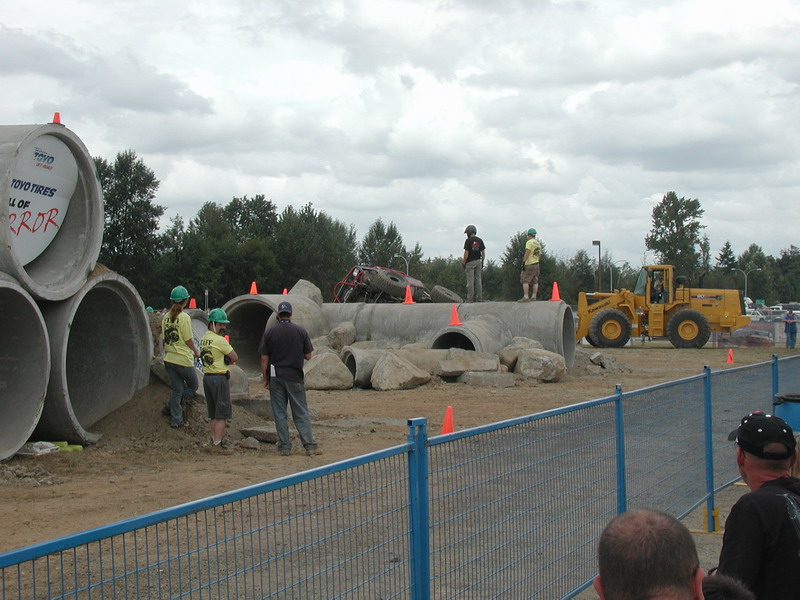 Red off-road vehicle on concrete pipes with yellow loader and observers.
