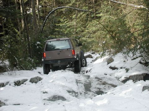 1985 Ford Bronco - A tan Ford Explorer is navigating a snowy, wooded trail with large rocks and a culvert visible.