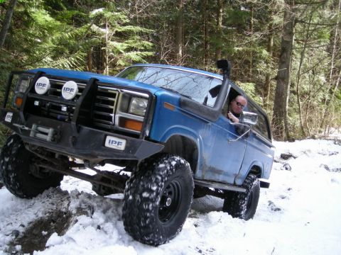 1985 Ford Bronco - A blue Ford Bronco with large off-road tires and a winch is navigating a snowy, wooded trail. The driver is visible through the open window…