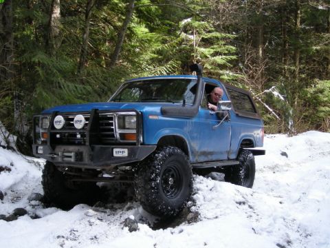 1985 Ford Bronco - A blue Ford Bronco is driven off-road through a snowy forest path. The vehicle features a snorkel, a brush guard with IPF lights, and large…