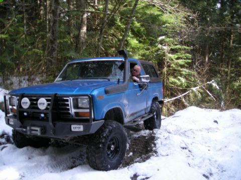 1985 Ford Bronco - A blue Ford Bronco with off-road modifications drives through a snowy, wooded area. The vehicle features a snorkel, a heavy-duty front bump…