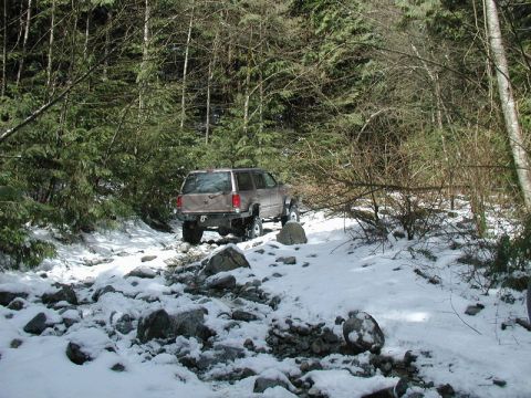 1985 Ford Bronco - A tan SUV navigates a rocky, snow-covered forest road with dense trees on either side.