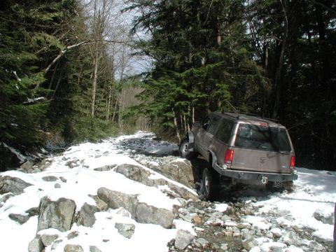 1985 Ford Bronco - A tan Ford Explorer is navigating a rocky, snow-covered trail through a dense forest.