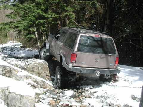 1985 Ford Bronco - A tan Ford Explorer is navigating a rocky and snowy terrain with trees in the background. The vehicle is angled to the right, showing its r…