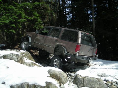 1985 Ford Bronco - A brown Ford Explorer is navigating a rocky, snow-covered trail with trees in the background. The vehicle is angled upwards, with its front…