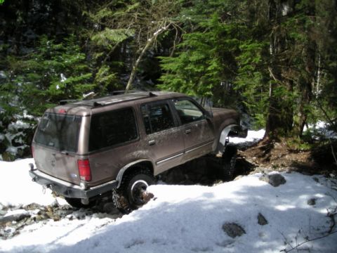 1985 Ford Bronco - A brown SUV is navigating a snowy, wooded trail, with one wheel deep in a rut and the vehicle tilted.