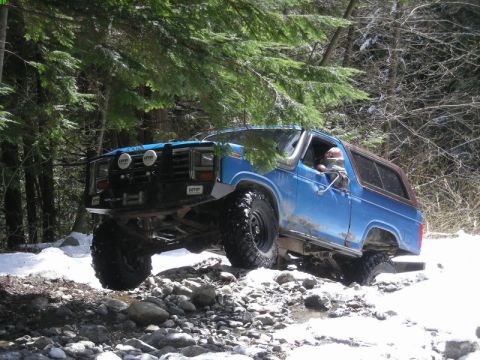 1985 Ford Bronco - A lifted blue 1980s Ford Bronco is navigating a rocky, snow-covered trail through a forest. The vehicle has large off-road tires, auxiliary…