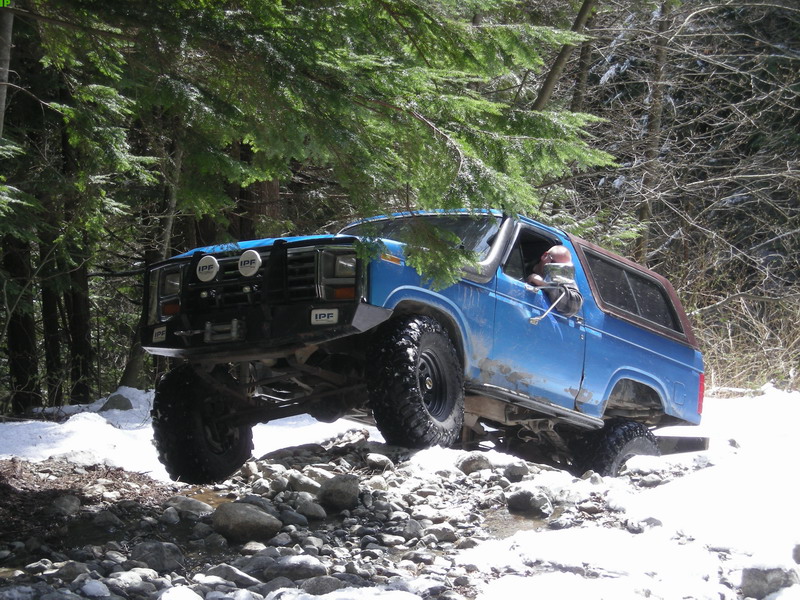 Lifted blue 1980s Ford Bronco off-roading on a rocky, snowy forest trail.