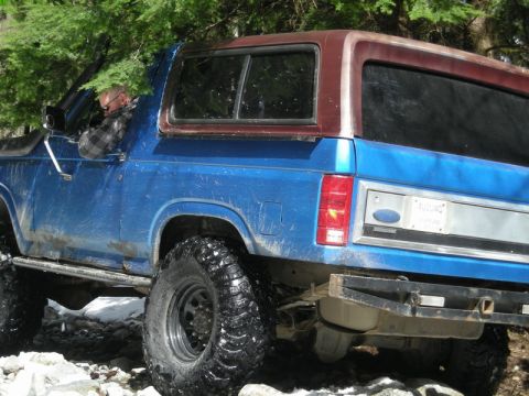1985 Ford Bronco - The rear driver's side of a blue Ford Bronco is shown off-road, navigating rocky terrain with large tires. A person wearing a plaid shirt i…
