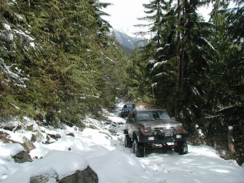 1985 Ford Bronco - A lifted, modified brown SUV drives down a snow-covered, rocky trail surrounded by evergreen trees. Another blue SUV is visible further up…