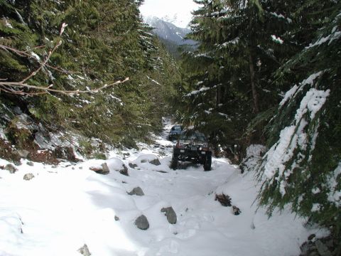 1985 Ford Bronco - Two off-road vehicles drive through a snowy, wooded mountain pass. The lead vehicle is a dark-colored SUV, possibly a Bronco, with large ti…