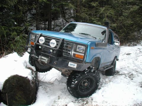 1985 Ford Bronco - A blue 1980s Ford Bronco is shown navigating a snowy forest trail, with its front tire resting on a large fallen log. The vehicle is equipp…