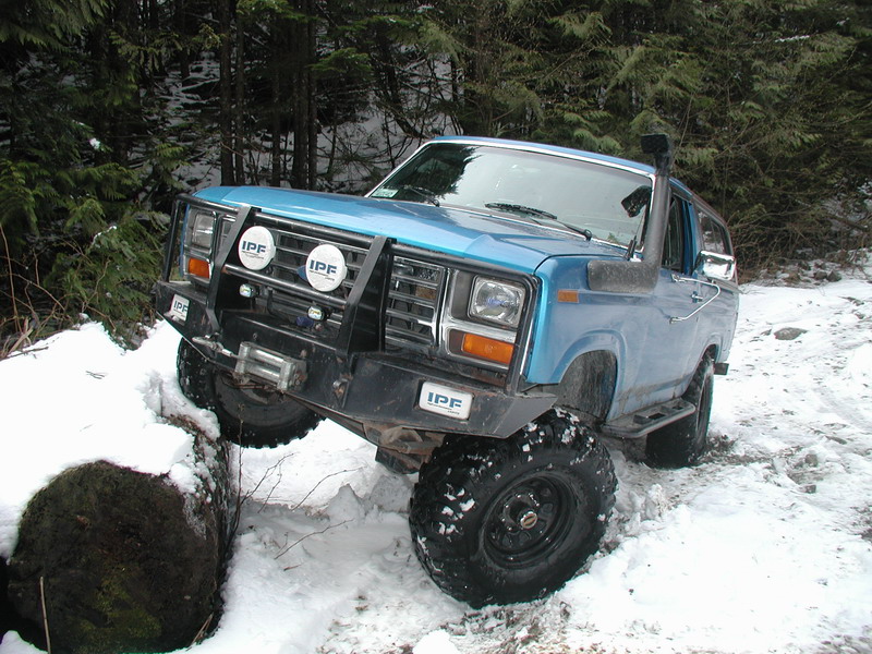 Blue 1980s Ford Bronco off-roading in snow, front tire on log, with winch and snorkel.