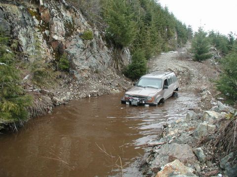 1985 Ford Bronco - A tan Ford Explorer drives through a muddy, shallow stream on a rocky, wooded trail. The vehicle has a winch mounted on the front bumper.