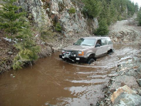 1985 Ford Bronco - A tan Ford Explorer SUV drives through a shallow, muddy creek bed with rocky, forested hillsides in the background. The vehicle has a winch…