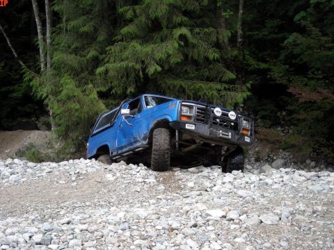 1985 Ford Bronco - A blue Ford Bronco is navigating a rocky, off-road trail with dense green trees in the background. The vehicle is angled upwards on a mound…