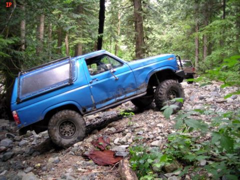 1985 Ford Bronco - A blue Ford Bronco is navigating a rocky, wooded trail, with its tires angled to climb over obstacles. Another off-road vehicle is visible…