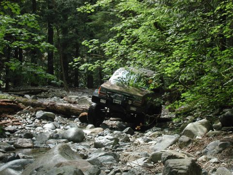 1985 Ford Bronco - A dark colored SUV is navigating a rocky stream bed surrounded by dense forest. The vehicle is angled towards the right, with its front end…