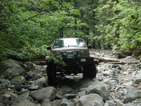 1985 Ford Bronco - A tan 1980s Ford Bronco is driving through a rocky, wooded stream bed. The vehicle has a modified front bumper and large off-road tires.