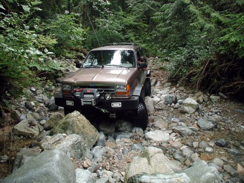 1985 Ford Bronco - A tan Ford Explorer is navigating a rocky, wooded stream bed. The SUV is equipped with an aftermarket bumper and a winch on the front.