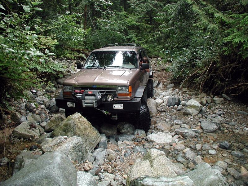 Tan Ford Explorer off-roading through a rocky stream in a forest.