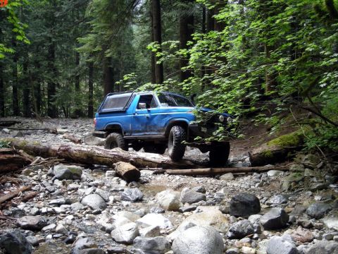 1985 Ford Bronco - A blue 1980s Ford Bronco with a cap is navigating a rocky, wooded terrain, driving over a fallen log in a dry creek bed.