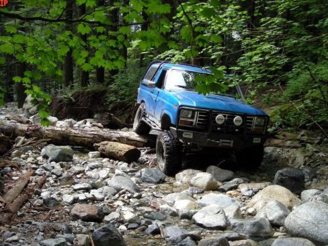 1985 Ford Bronco - A blue 1980s Ford Bronco is navigating a rocky stream bed in a forest. The vehicle is angled towards the viewer, showing its front grille,…