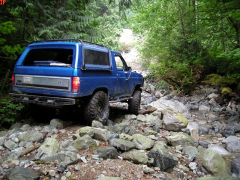 1985 Ford Bronco - A blue Ford Bronco is navigating a rocky, wooded trail next to a stream. The vehicle is viewed from the rear-side angle, showcasing its off…